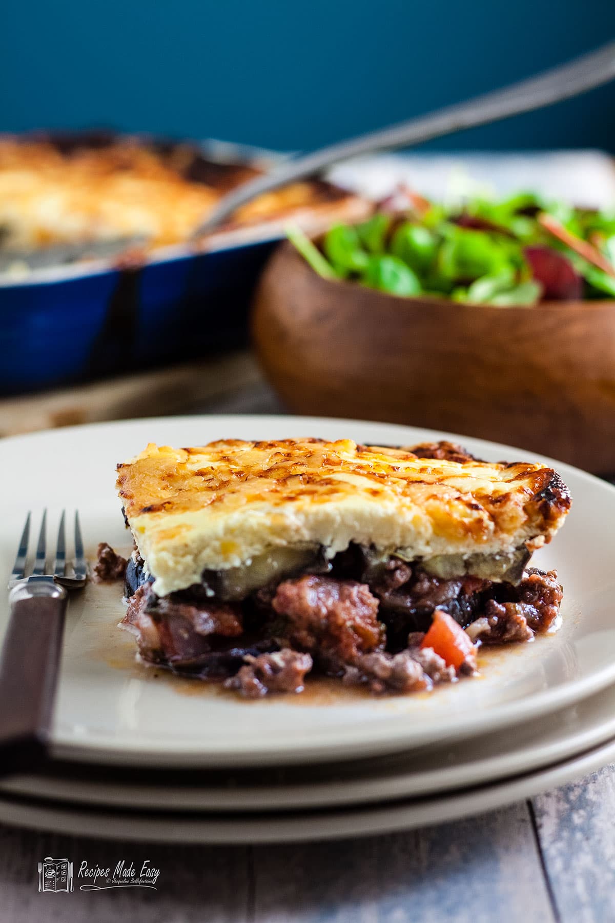 portion of moussaka on a plate with bowl of salad and serving dish behind.