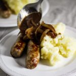 spooning gravy over sausages and mash in a plate.