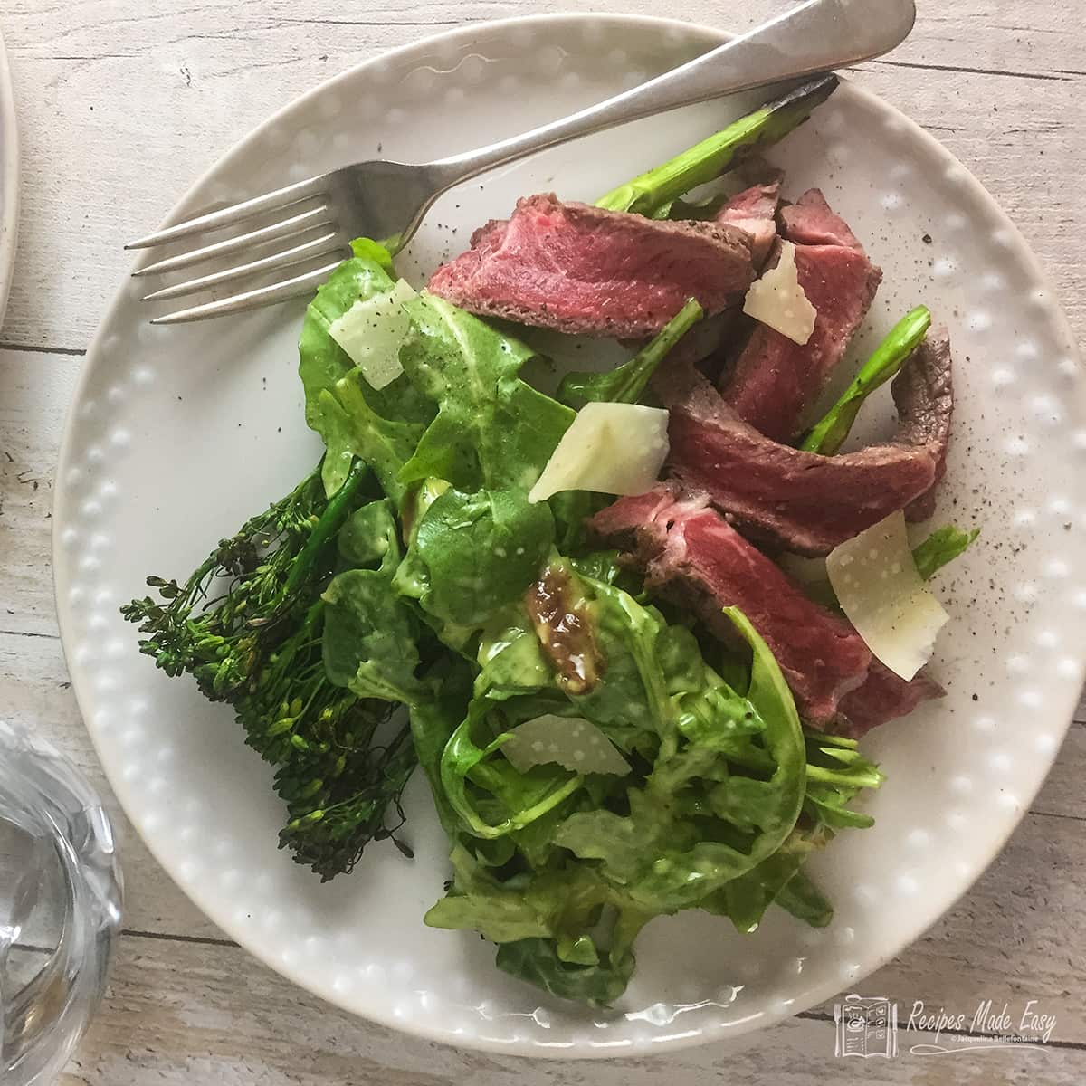 chargrilled broccoli rocket and beef salad on dinner plate with glasses behind. dress