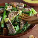 Serving bowl of chicken Caesar salad with asparagus. Bowl of Caesar salad dressing behind.