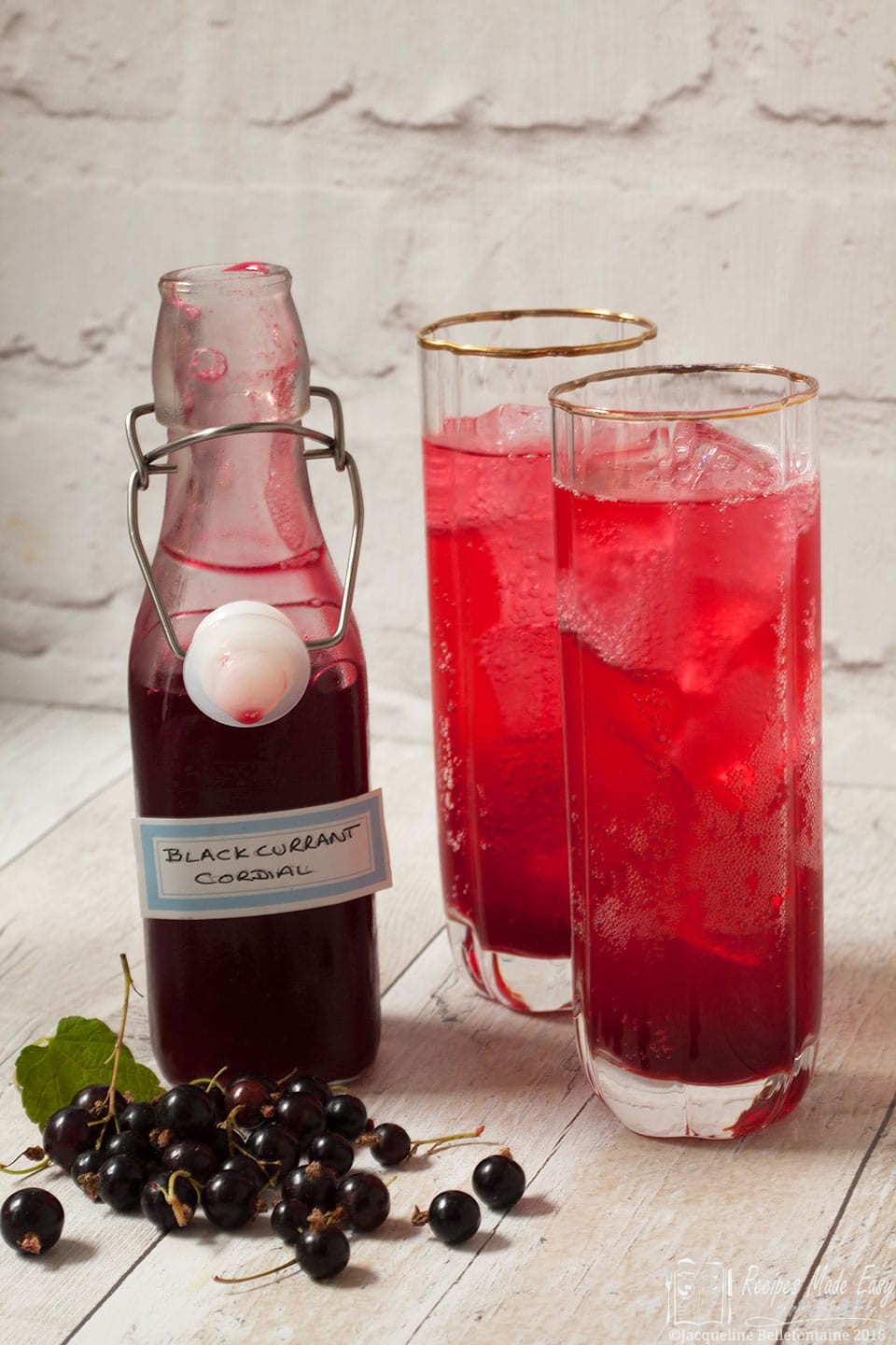Bottle of homemade blackcurrant cordial by recipes made easy with two glasses of cordial to the side.