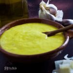 bowl of Caesar salad dressing with wooden spoon. parmesan cheese in front and garlic behind.