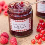 Open jar of raspebrry and redcurrant jam by recipes made easy. Spoon in jar and surrounded by raspberries and redcurrants.