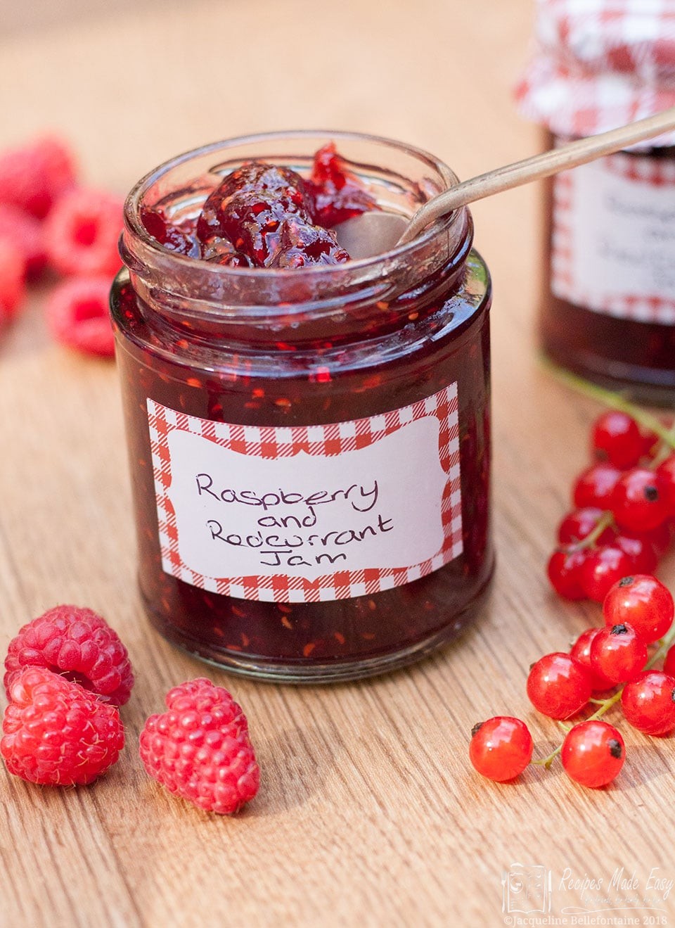 Open jar of raspebrry and redcurrant jam by recipes made easy. Spoon in jar and surrounded by raspberries and redcurrants.