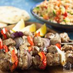 easy lamb kebabs by recipes made easy on a wooden tray with lemon wedges as garnish and salad , flatbreads and sauce in background