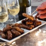 Party sausages on dish with hand reaching in to take one on a cocktail stick. Glasses behind.