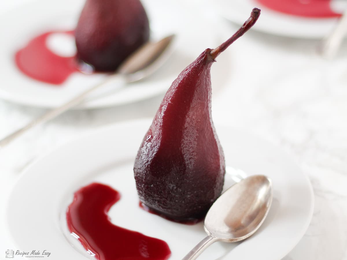 poached pear standing on a white plate with pool of syrup and a teaspoon. Two more serving behind.