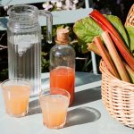 basket of rhubarb on table with bottle of corial, jug of water and two poured glasses of rhubarb cordial