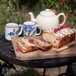 raspberry and white chcolate loaf cake on a wooden board with teapot and mugs in background