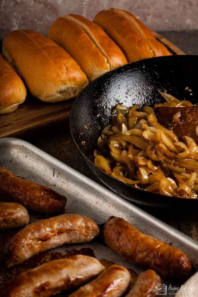 tray of sausages plus onions in a pan and bread rolls behind