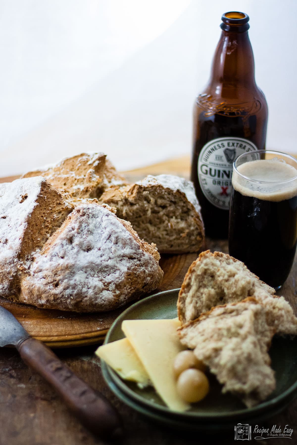 Irish soda bread loaf on a board with portion on plate served with cheese and pickles.  Bottle and glass of guinness behind.