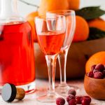 bottle of cranberry and clementine gin with some poured into a glass in front of bowl of clementines.