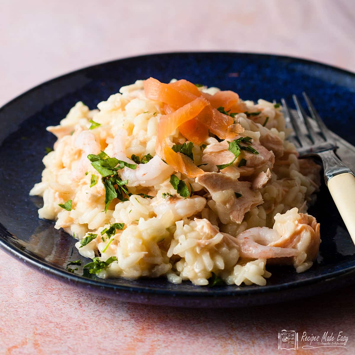 Portion of salmon and prawn risotto on a blue plate with knife and fork on the side.