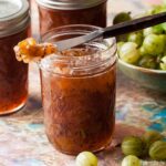 open jar of easy gooseberry jam with some on a knife resting on top. Unopened jars a bowl of gooseberries behind.