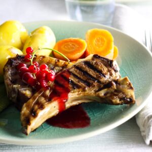 2 lamb chop on a plate with redcurrant jus on top garnished with a sprig of fresh redcurrants and served with potatoes and carrots.