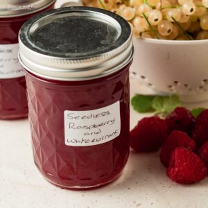 Jar of raspberry and whitecurrant jam with a colander of white currants behind and a few raspberries to the side.